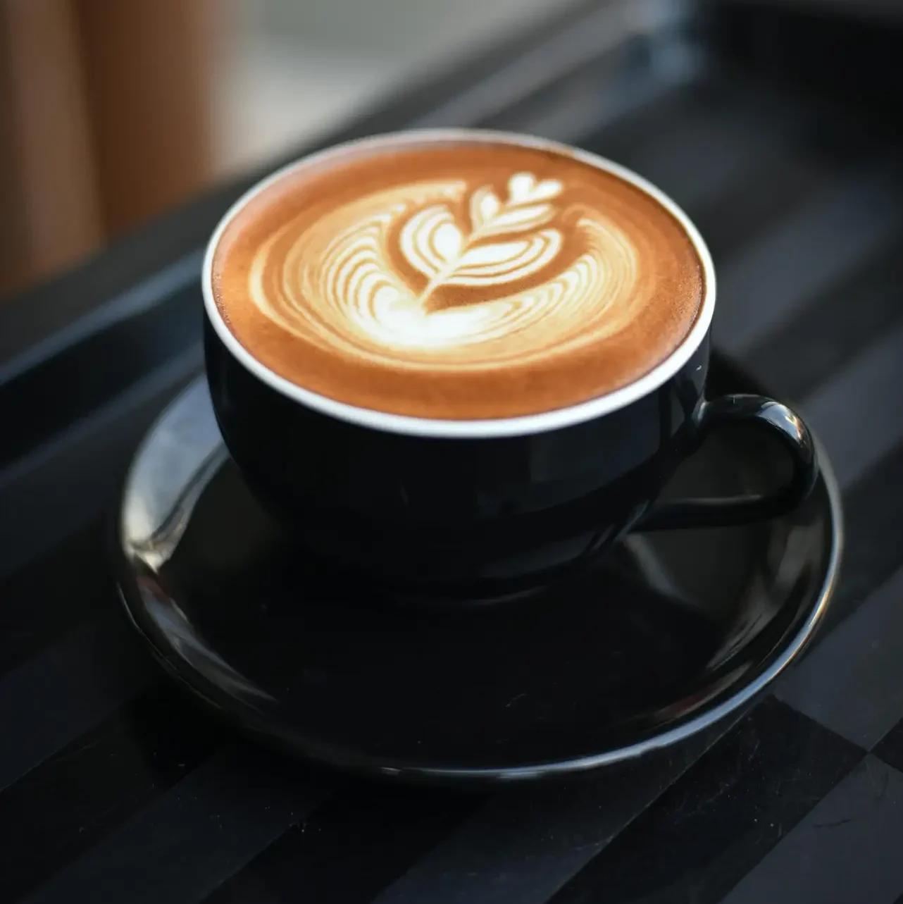 A beautifully crafted cappuccino with intricate latte art in a black ceramic mug on a dark surface.